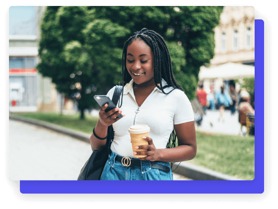 Young woman smiling while looking at her phone and holding a coffee cup outdoors.