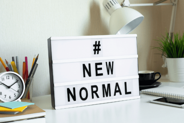 A desk with a lightbox reading "#NEW NORMAL," surrounded by stationery, a clock, and a coffee cup.