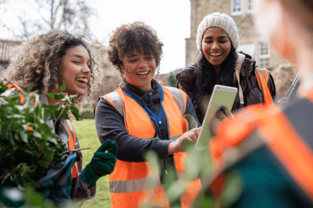 Three women in safety vests smile while looking at a smartphone during an outdoor community activity.