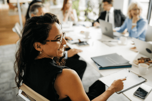 The image shows a smiling woman wearing glasses and sitting at a conference table with other colleagues.