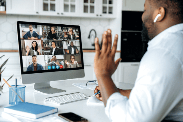 A man in earbuds waves during a virtual call with nine participants, indicating a remote work environment.