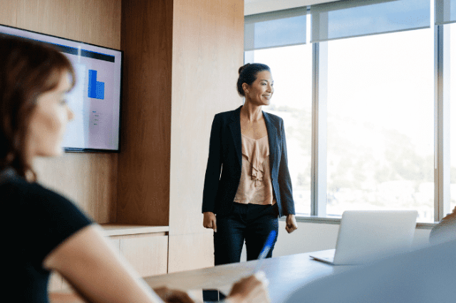 A woman stands confidently, presenting to colleagues in a conference room with a chart displayed on the screen.