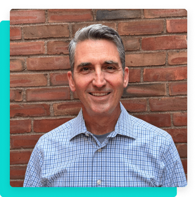 Portrait of a smiling man, Chip Corboy, standing in front of a brick wall.