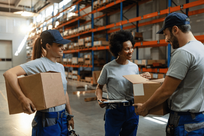 Three people in gray shirts and navy blue hats holding boxes in a warehouse.