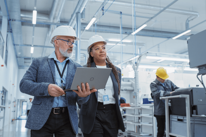 Two people in white hard hats walking a manufacturing plant floor.