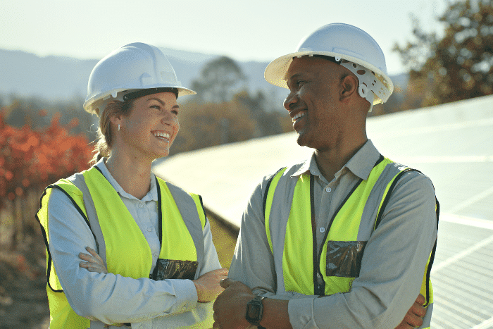 A man and woman talking. They are outside, wearing white hard hats and safety vests, in front of solar panels.