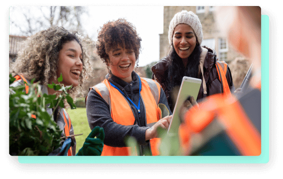 Three women in safety vests smile while looking at a smartphone during an outdoor community activity.