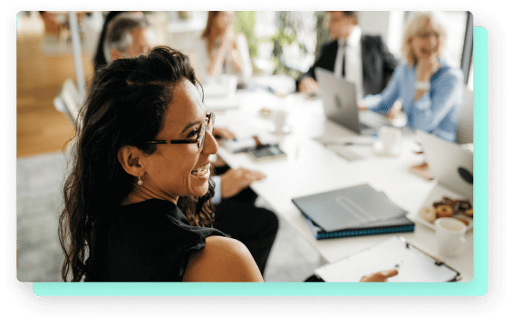 The image shows a smiling woman wearing glasses and sitting at a conference table with other colleagues.