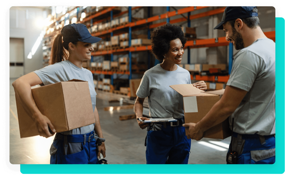 Three people in gray shirts and navy blue hats holding boxes in a warehouse.